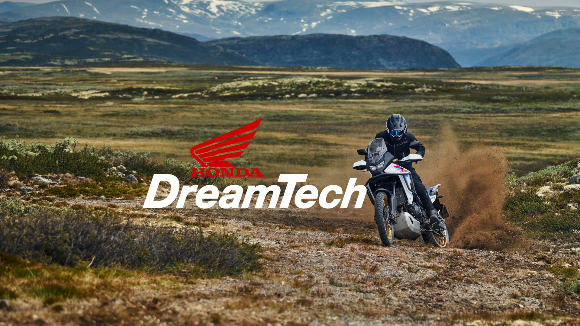 A Honda motorcycle rider traverses a dusty trail in a vast, mountainous landscape. The image features the DreamTech logo, which includes Honda's iconic red wing symbol, overlaid on the scenic background.